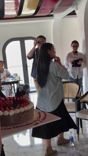 Professional video production of an intimate celebration event at a modern Đà Lạt venue, featuring a decorated cake and group gathering in a bright, minimalist café space with professional lighting setup.